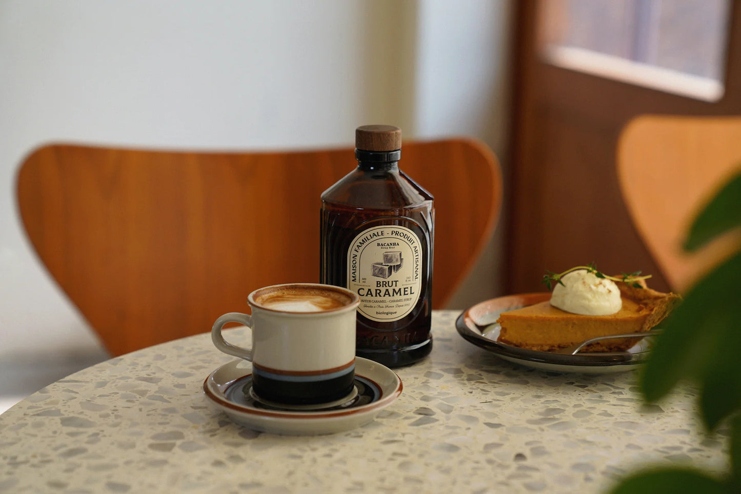 Bacanha Luomu Karamellisiirappi; latte valkoisessa posliinikupissa ja -aluslautasessa, tumma lasipullo Brut Caramel -etiketillä lihavoidulla serif-fontilla, sekä kermalla koristeltu kolmionmuotoinen piirakanpala pyöreällä valkoisella lautasella; asetelma laikukkaalla terrazzo-pöydällä; taustalla puutuoli ja ovi, vihreä kasvi etualalla.