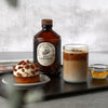 Bacanha Luomu Karamellisiirappi; karamellisiirappipullo, jäillä täytetty iced latte kirkkaassa lasissa, pekaanijälkiruoka valkoisella lautasella ja pieni siirappiannos aseteltuna suorakaiteen muotoiselle mustalle mattapintaiselle tarjottimelle; siirappipullossa beige etiketti mustalla groteskitekstillä; taustalla lehti pöydällä; ei ihmisiä tai käsiä.