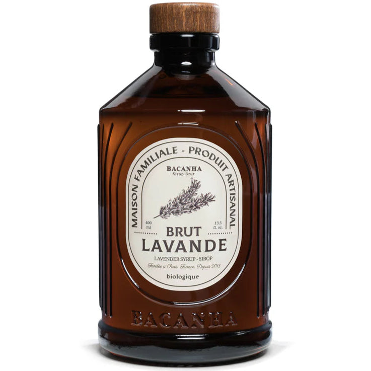 Bacanha Luomu Laventelisiirappi; ruskea lasipullo puisella korkilla, merkitty Brut Lavande -laventelisiirappi, valmistaja Bacanha; kermanvärisessä etiketissä mustaa antiikvatekstiä, yksityiskohtainen laventelikuvitus ja hienoa kasviaiheista viivapiirrosta; pullo on sileä, kiiltäväpintainen ja sylinterinmuotoinen; kuvattu valkoista taustaa vasten tasaisessa valaistuksessa, ei ihmisiä tai käsiä näkyvissä.