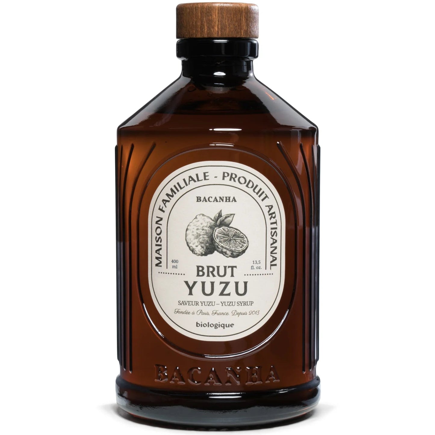 Bacanha Luomu Yuzusiirappi; ruskea lasipullo Bacanha Brut Yuzu -luomusiirappia mustalla muovikorkilla, sylinterinmuotoinen ja kiiltäväpintainen; valkoisessa etupuolen etiketissä keltainen yuzuhedelmäkuvitus ja ranskankielistä tekstiä lihavalla groteskilla ja antiikvalla; tausta valkoinen; tuote esillä pystyssä, ei ihmisiä tai käsiä kuvassa.
