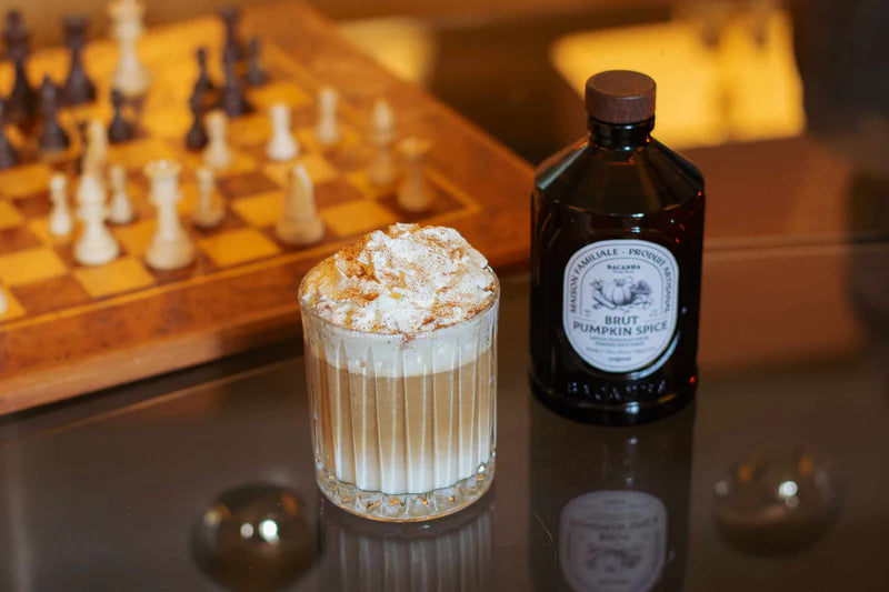 Bacanha Pumpkin Spice Siirappi; kirkas lasi täynnä vaaleanruskeaa, vaahtoavaa lattea kermavaahdolla ja kanelilla, aseteltuna lieriömäisen pullon viereen, jonka etiketissä lukee Brut Pumpkin Spice lihavoiduin isoin groteskikirjaimin; molemmat läpinäkyvällä lasipöydällä, taustalla puinen shakkilauta; ei ihmisiä tai käsiä.