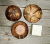 Two round coconut shell soap dishes, one with drainage holes and the other holding a pink soap, rest alongside a white bar of soap on a wooden surface. Surfaces of the dishes are matte with visible natural texture and color variations typical of coconut shells. No packaging or labels are present. Background is an unvarnished wood tabletop; no people or hands are visible in the frame.