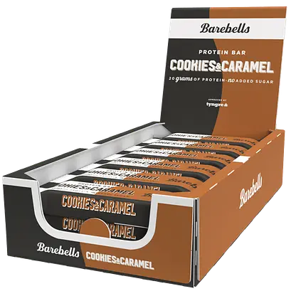 Barebells Cookies-Caramel Proteiinipatukka; laatikko Barebells-proteiinipatukoita, maku cookies & caramel; pakkaus on suorakulmainen mattamustalla pohjalla, ruskein yksityiskohdin ja valkoisin lisäelementein; patukat on aseteltu riveihin avonaisen laatikon sisällä; laatikossa isokokoista groteskitekstiä ”20 grams of protein” ja ”no added sugar”; ei ihmisiä tai käsiä näkyvissä; tausta on yksivärinen ja pinnaltaan tasainen.