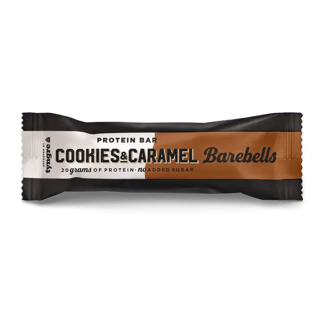 Barebells Cookies-Caramel Proteiinipatukka; Barebells-proteiinipatukka Cookies & Caramel -maussa, käärittynä valkoiseen, mustaan ja ruskeaan pakkaukseen mattapinnalla; suorakulmainen muoto ryppyisin reunoin; typografia lihavaa ja groteskia, ”20 grams of protein” ja ”no added sugar” näkyvästi kääreessä; ei ihmisiä tai käsiä näkyvissä; tausta vaalea ja pehmeästi valaistu, tuote selkeästi rajattuna; herkullinen välipala aktiiviseen elämäntapaan.