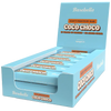 Barebells Soft Coco Choco Proteiinipatukka; sininen suorakaiteen muotoinen näyttölaatikko, jossa useita yksittäin käärittyjä Barebells Coco Choco -pehmeitä proteiinipatukoita; kääreet ja laatikko sinisiä, valkoista ja ruskeaa tekstiä; pakkauksessa lukee 16 grammaa proteiinia ja ei lisättyä sokeria selkeällä groteskifontilla; pinta mattapintainen; ei ihmisiä tai rekvisiittaa; tausta vaalea.