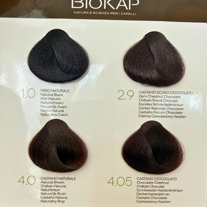 Biokap Nutricolor Delicato Kestoväri; neljä hiusvärinäytettä merkinnöin: 1.0 Natural Black, 2.9 Dark Chestnut Chocolate, 4.0 Natural Chestnut ja 4.05 Chocolate Chestnut näytetään Biokap-hiusvärikartassa vaakarivissä; jokainen näyte kiinnitetty suorakulmaiseen valkoiseen korttiin painetuin mustin etiketein ja brändinimellä groteskikirjasimella; ei ihmisiä tai käsiä kuvassa, tausta valkoinen.