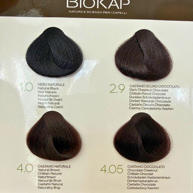 Biokap Nutricolor Delicato Kestoväri; neljä hiusvärinäytettä merkinnöin: 1.0 Natural Black, 2.9 Dark Chestnut Chocolate, 4.0 Natural Chestnut ja 4.05 Chocolate Chestnut näytetään Biokap-hiusvärikartassa vaakarivissä; jokainen näyte kiinnitetty suorakulmaiseen valkoiseen korttiin painetuin mustin etiketein ja brändinimellä groteskikirjasimella; ei ihmisiä tai käsiä kuvassa, tausta valkoinen.