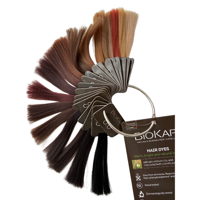 Biokap Nutricolor Delicato Kestoväri; viuhka hiusvärinäytteitä eri sävyissä vaaleasta vaaleasta tummanruskeaan, kiinnitettynä metallirenkaaseen tuotetietokortin kera merkinnällä BIOKAP innovation; säikeet ryhmitelty rinnakkain tasaiseen riviin, kukin erillinen mutta yhdistetty yläreunasta; näytteet vaikuttavat suorilta ja kiiltäviltä; kortti suorakulmainen yksinkertaisella tekstityylillä; neutraali tausta, lähikuva tuotteesta, ei ihmisiä tai käsiä.