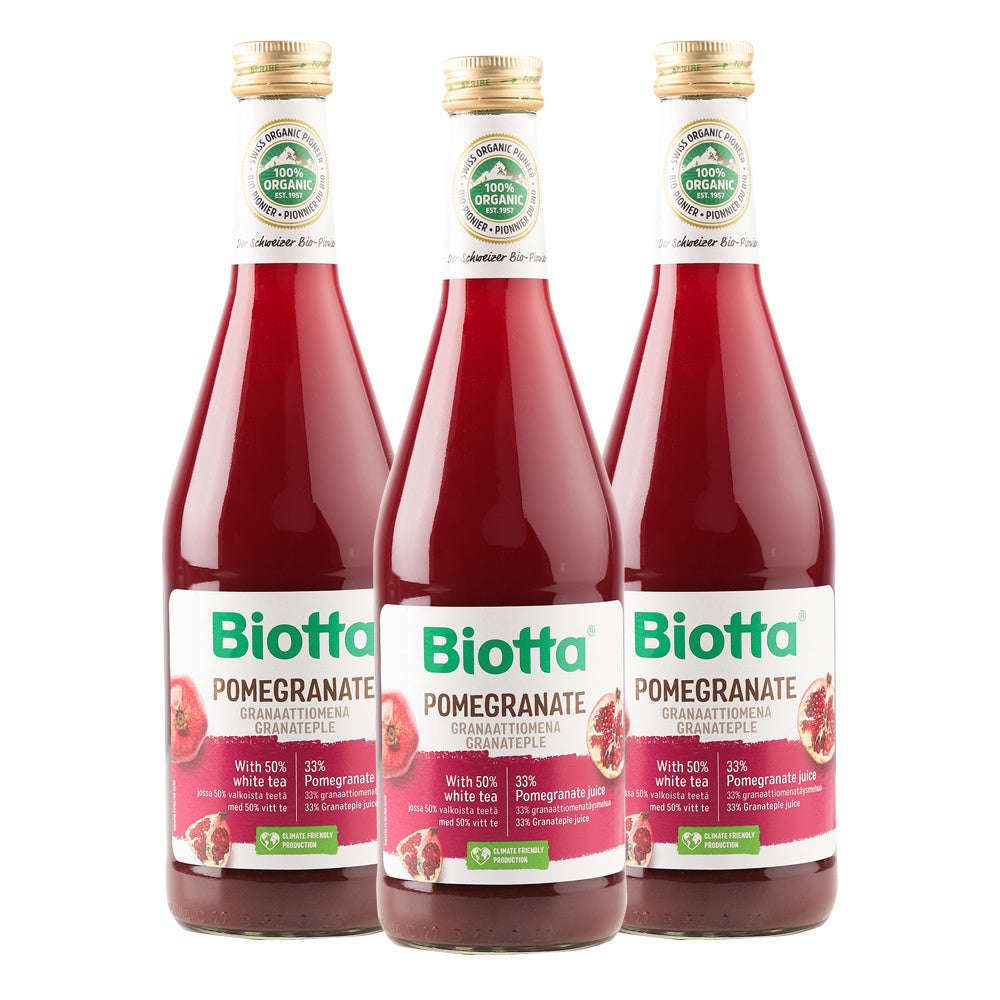 Three glass bottles of Biotta Pomegranate juice are shown side by side. The transparent bottles have a slight taper toward the neck, finished with metallic gold screw caps. Labels are green and white with glossy surfaces, showing a pomegranate image and bold sans-serif “Biotta” text. Background and setting are neutral and unadorned. No people or hands are present in the image.