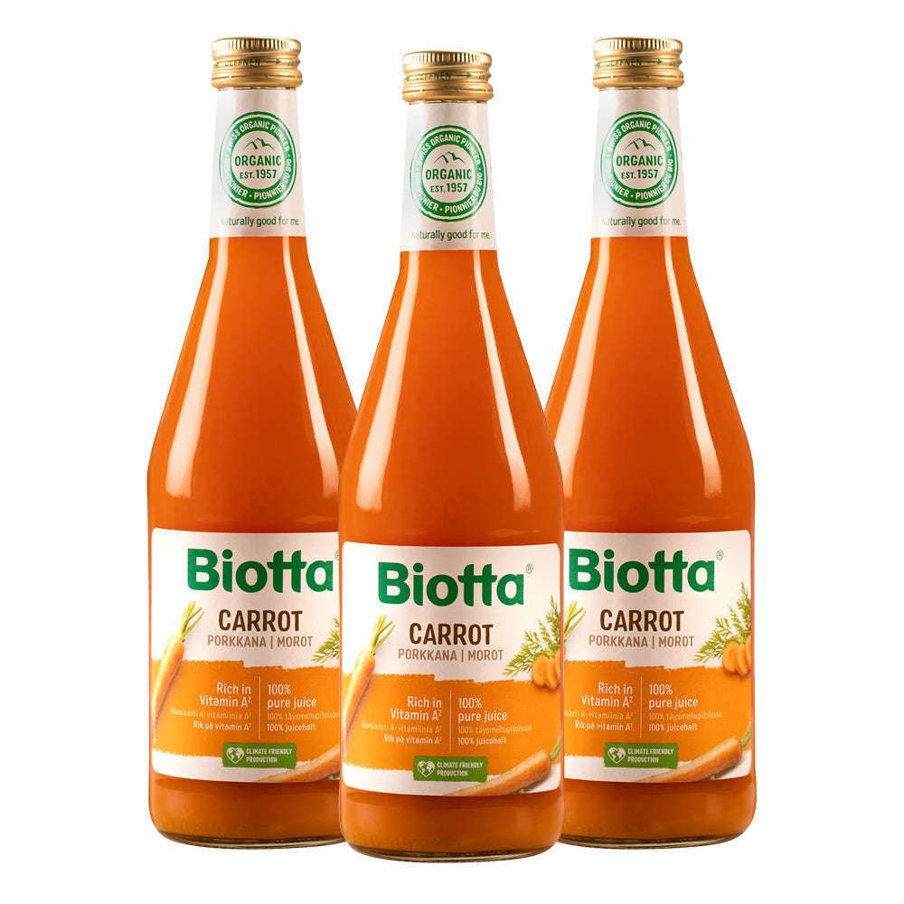 Three glass bottles of Biotta Carrot Juice, labeled as organic and rich in vitamin A, stand side by side. Labels display orange carrot graphics, green sections with white text, and the Biotta logo in bold sans-serif lettering. Each bottle has a glossy transparent surface, tapered shape, and is topped with a white screw cap featuring green and silver seals. The background is plain white. No people are visible.