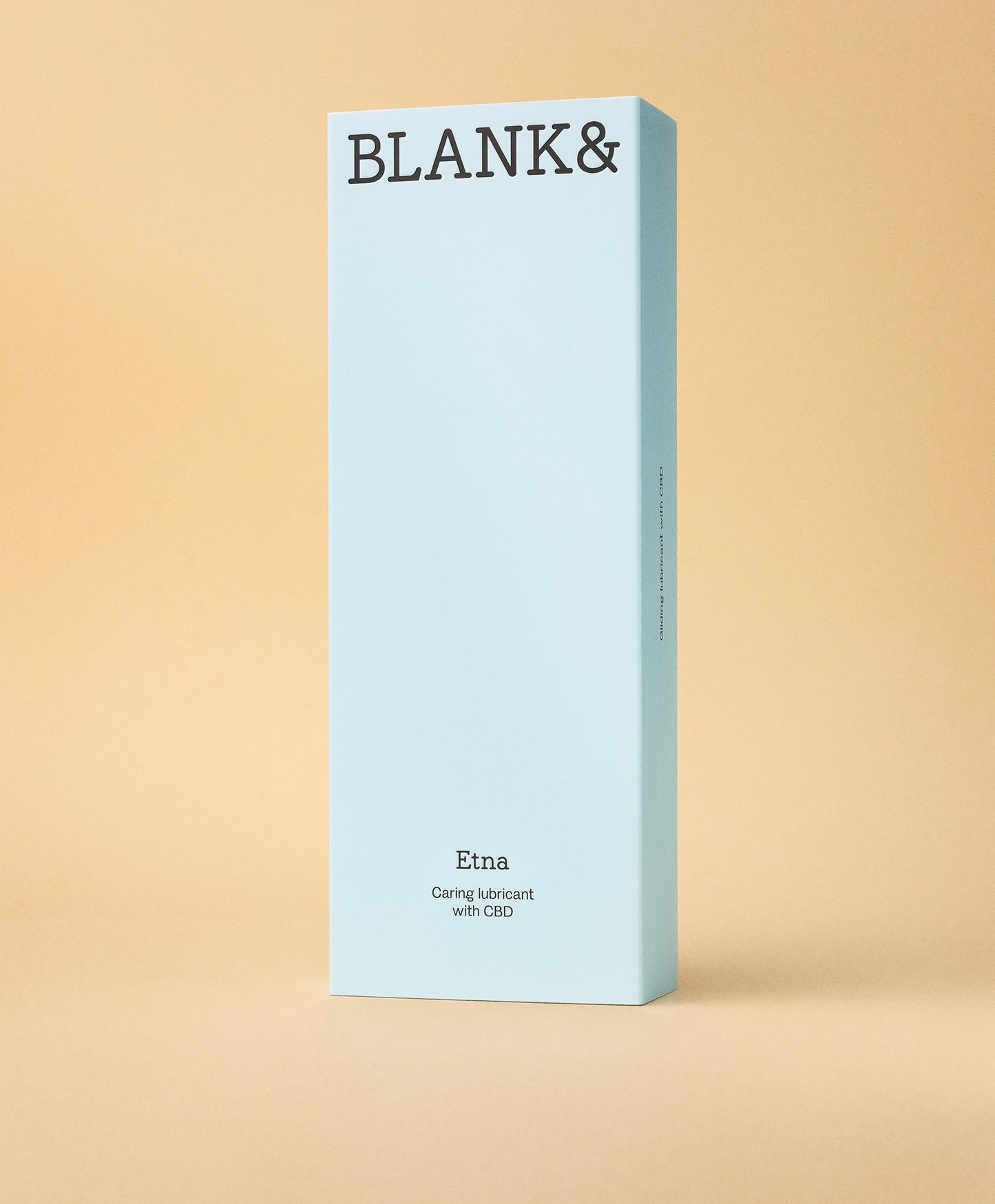 Blank& Etna Liukuvoide; suorakulmainen vaaleansininen rasia tekstillä BLANK& yläosassa seisoo pystyssä tasaista beigeä taustaa vasten; rasia matalla pinnalla, kuvattu suoraan edestä; pakkaus pahvia; ainoa luettava teksti kuuluu ”Etna Caring lubricant with CBD”; ei ihmisiä tai muita esineitä kuvassa.