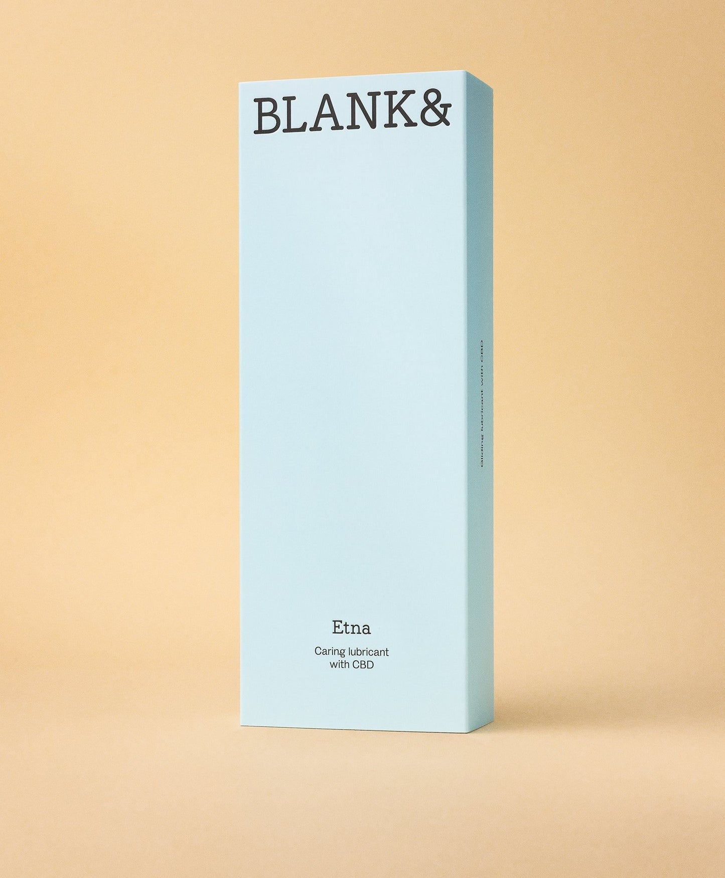 Blank& Etna Liukuvoide; suorakulmainen vaaleansininen rasia tekstillä BLANK& yläosassa seisoo pystyssä tasaista beigeä taustaa vasten; rasia matalla pinnalla, kuvattu suoraan edestä; pakkaus pahvia; ainoa luettava teksti kuuluu ”Etna Caring lubricant with CBD”; ei ihmisiä tai muita esineitä kuvassa.