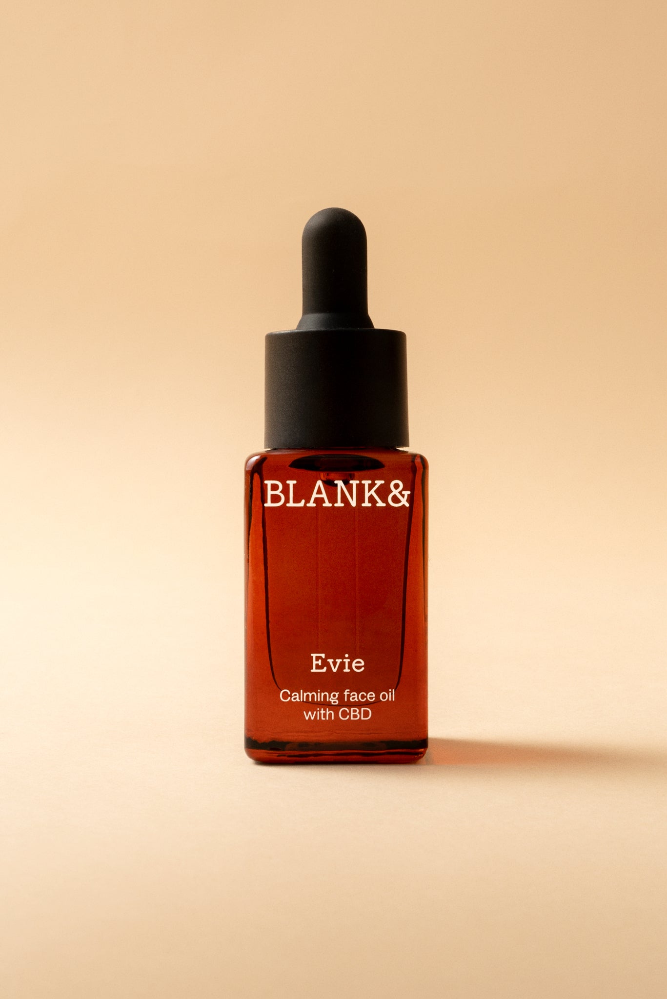 Blank& Evie Kasvoöljy; ruskea lasinen tippapullo tekstillä ”BLANK& Evie Calming face oil with CBD” seisoo pystyssä vaaleaa beigeä taustaa vasten; pullo sylinterin muotoinen pyöristetyin olkapäin ja mustalla tippakorkilla; etikettiteksti selkeää, mustaa groteskikirjasinta; lasin pinta kiiltävä ilman näkyvää kuviointia; ei ihmisiä tai käsiä kuvassa.