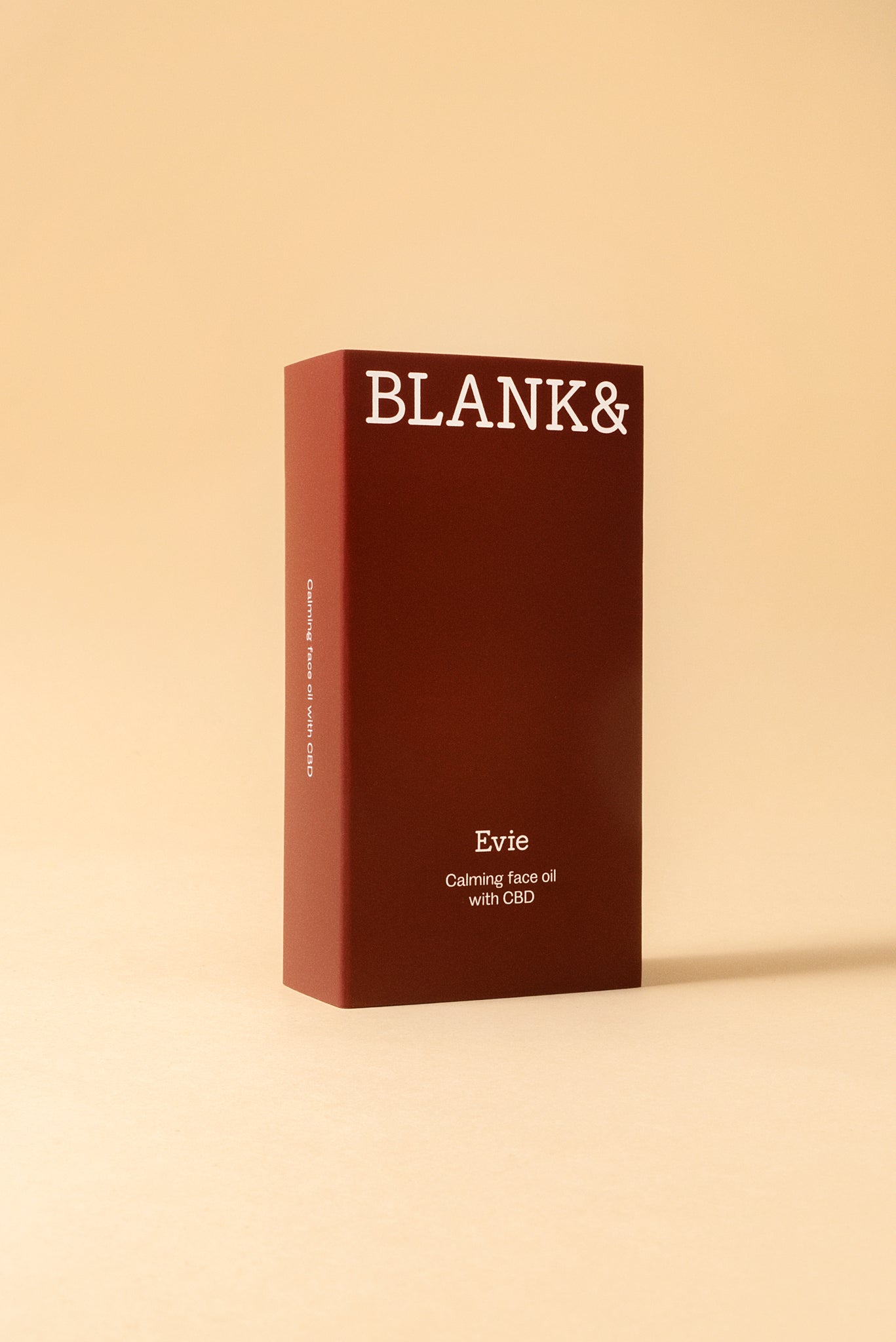 Blank& Evie Kasvoöljy; punainen suorakulmainen mattapintainen rasia näyttää tekstin ”BLANK&” yläosassa ja ”Evie Calming face oil with CBD” alaosassa selkeällä nykyaikaisella groteskikirjasimella; tuote kuvattu tasaista beigeä taustaa vasten; ei ihmisiä tai käsiä kuvassa; asetelma välittää rauhallisen tunnelman.