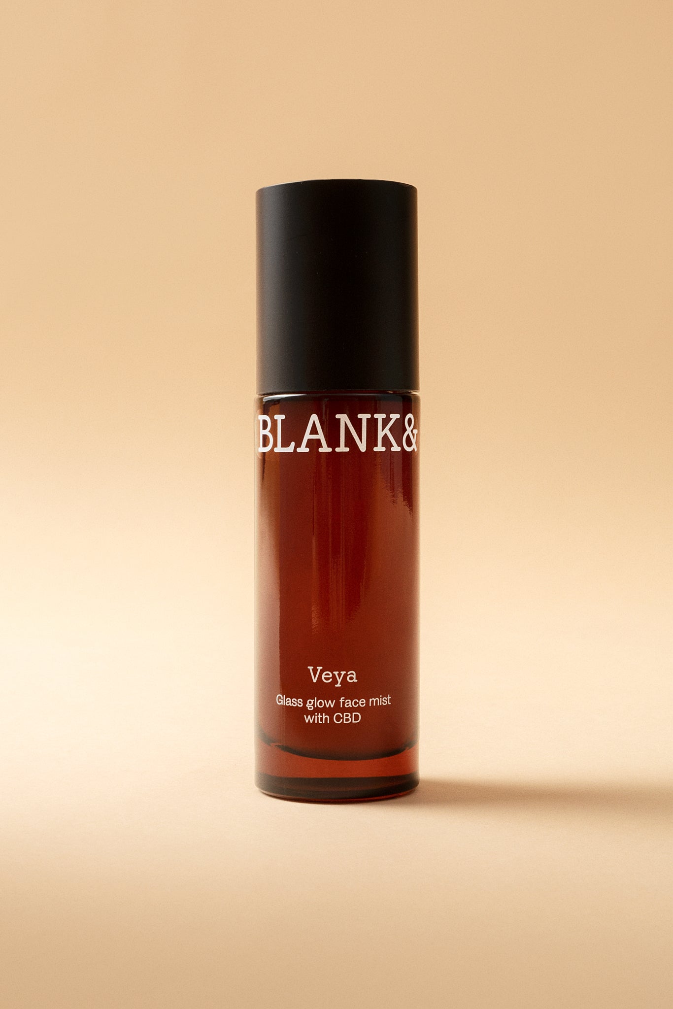 Blank& Veya Kasvosuihke; ruskea pullo mustalla korkilla tekstillä BLANK& ja Veya Glass glow face mist with CBD and hemp seisoo pystyssä beigeä taustaa vasten; pullo sylinterin muotoinen, todennäköisesti lasia, kiiltopinnalla; etiketissä yksinkertaista mustaa groteskitekstiä valkoisella pohjalla; ei ihmisiä tai käsiä kuvassa; kuva otettu edestä silmän korkeudelta tasaisella taustalla.