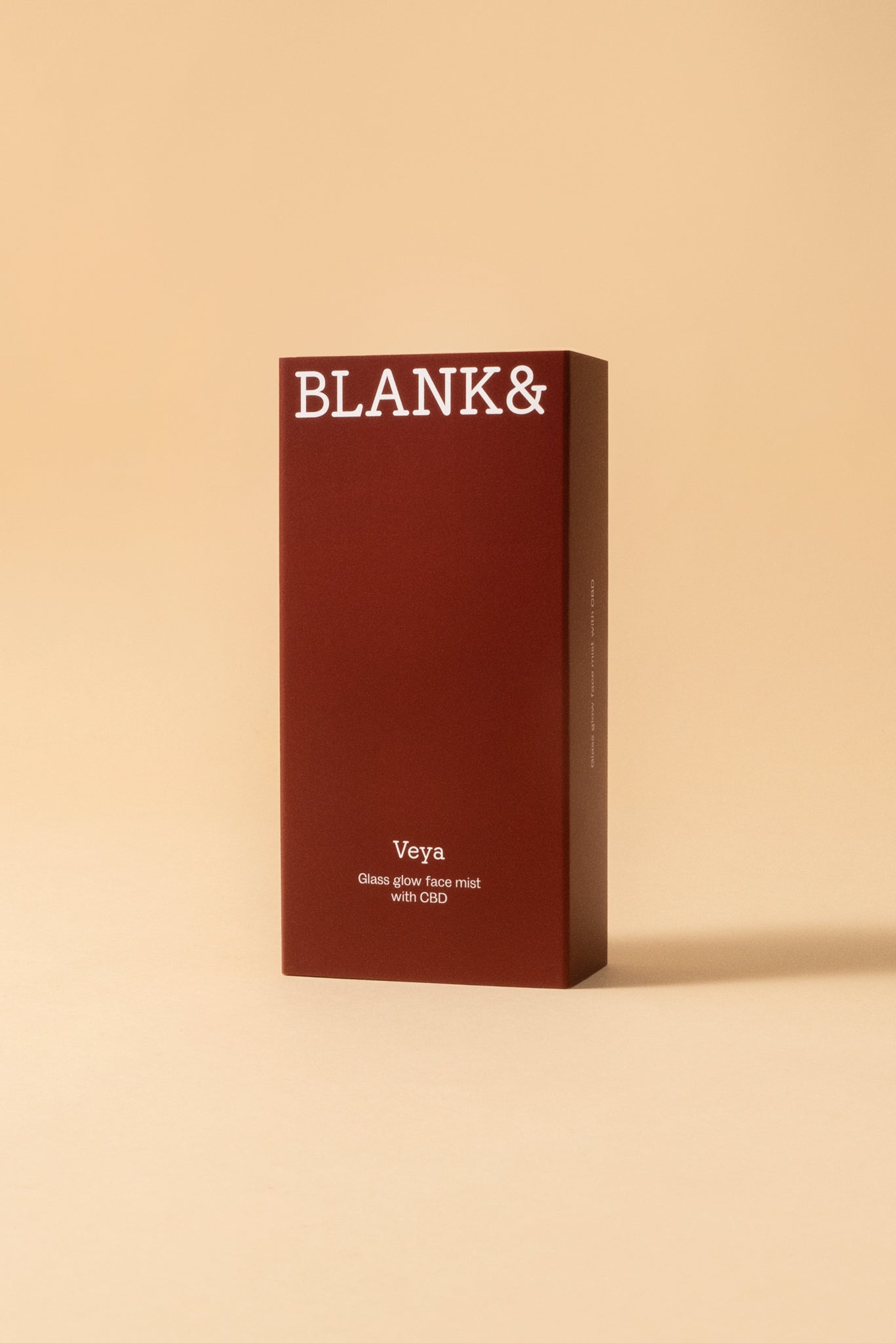 Blank& Veya Kasvosuihke; tummanpunainen suorakulmainen rasia tekstillä BLANK& yläosassa, alla teksti Veya Glass Glow Face Mist with CBD ja maininta säteilevästä lopputuloksesta, vaaleaa beigeä taustaa vasten; rasian pinta sileä ja kulmat terävät; teksti selkeällä nykyaikaisella groteskikirjasimella; ei ihmisiä tai käsiä, kuva otettu edestä tasaisessa valaistuksessa.