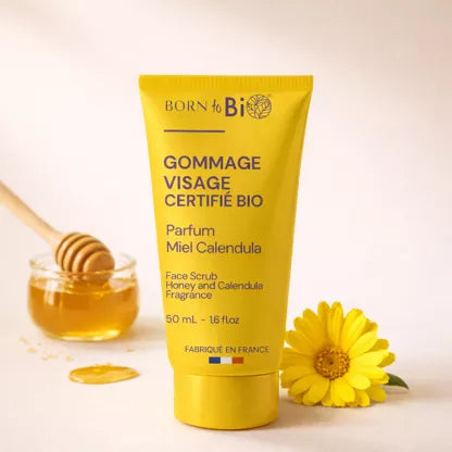 Born to Bio Face Scrub Honey & Calendula: keltainen tuubi tekstillä "Born to Bio Face Scrub" lihavoidulla, selkeällä kirjasimella pystyasennossa; tuubi on mattapintainen ja siinä yksinkertaisia graafisia elementtejä; vieressä läpinäkyvä lasikulho hunajaa, puinen hunajakauha ja yksinäinen keltainen päivänkakkaramainen kukka; kaikki esineet ovat vaalealla, yksivärisellä alustalla; ei ihmisiä tai käsiä näkyvissä.