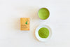 Clearspring Luomu Matcha-teejauhe Ceremonial Grade: rasia Clearspring-luomu japanilaista matchaa vihreä-valkoisella mattapakkauksella ja lihavoidulla mustalla typografialla seisoo pystyssä; vieressä pyöreä valkoinen kulho vihreää nestettä ja pieni valkoinen lautanen hienoa vihreää teejauhetta; kaikki esineet on aseteltu kirkkaalle, vaakasuoralle valkoiselle puupinnalle ylhäältä kuvattuna.