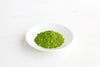 Clearspring Luomu Matcha-teejauhe Ceremonial Grade: pieni valkoinen lautanen, jolla kasa eloisan vihreää matchajauhetta, valkoisella pinnalla, näyttäen hienon tekstuurin ja rikkaan värin; lautanen on keraaminen, pyöreä, kiiltäväpintainen ja koristelematon; pakkausta tai tekstiä ei näy; tausta on valkoinen; kuva on lähikuva tuotteesta ilman muita esineitä tai ihmisiä.