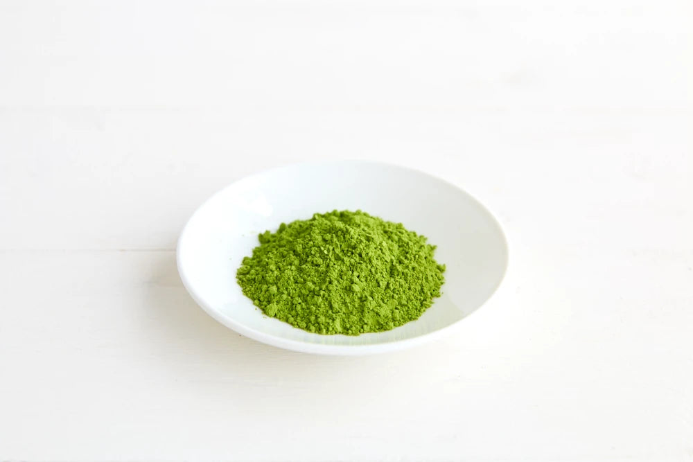 Clearspring Luomu Matcha-teejauhe Ceremonial Grade: pieni valkoinen lautanen, jolla kasa eloisan vihreää matchajauhetta, valkoisella pinnalla, näyttäen hienon tekstuurin ja rikkaan värin; lautanen on keraaminen, pyöreä, kiiltäväpintainen ja koristelematon; pakkausta tai tekstiä ei näy; tausta on valkoinen; kuva on lähikuva tuotteesta ilman muita esineitä tai ihmisiä.