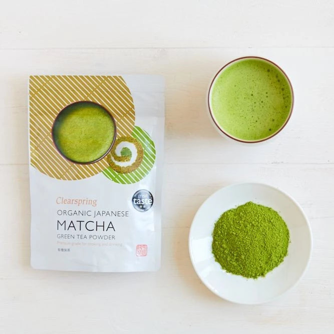 Clearspring Luomu Matcha-teejauhe Premium Grade: pussi Clearspring-luomu japanilaista matchajauhetta mattahopeisella ja vihreällä pakkauksella, selkeä tuote-etiketti, seisoo pystyssä tasaista valkoista taustaa vasten; vieressä pyöreä kulho kirkkaan vihreää valmista matchateetä, keraamista materiaalia kiiltäväpinnalla; pieni valkoinen lautanen sisältää kasan hienoksi jauhettua vihreää seremoniallista matchajauhetta; ei ihmisiä tai käsiä näkyvissä kuvassa.