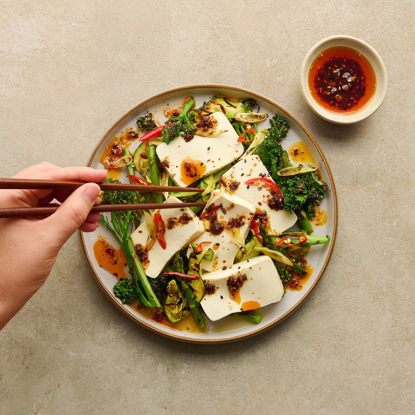 Clearspring Luomu Silken Tofu: käsi käyttää syömäpuikkoja poimimaan tofua pyöreältä valkoiselta lautaselta, jolla viipaloitua tofua, parsakaalinversoja ja sekoitettuja vihanneksia punaisella chilikastikkeella valeltuna; lautanen on beigellä sileällä pinnalla; pienessä keraamisessa kulhossa chiliöljyä lautasen vieressä; ei näkyviä etikettejä tai pakkausta kuvassa.