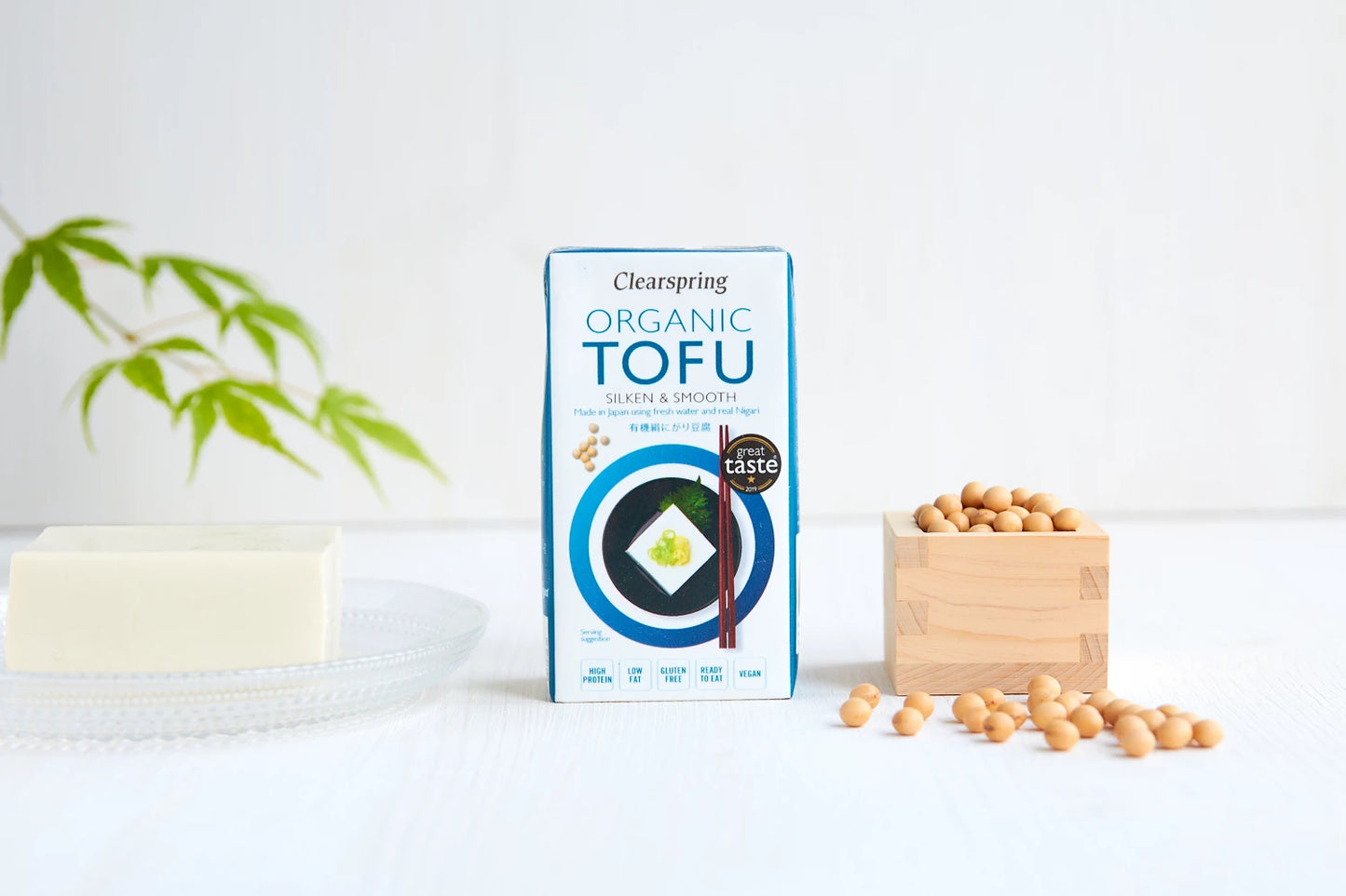 Clearspring Luomu Silken Tofu: rasia Clearspring-luomutofua seisoo pystyssä; pakkaus on valkoinen sini-mustalla groteskitekstillä; vasemmalla läpinäkyvä lasilautanen, jolla suorakulmaisia paloja sileää, vaaleaa silkkitofua; oikealla avoin, neliönmuotoinen puinen astia täynnä pyöreitä, vaaleita soijapapuja; kaikki esineet on aseteltu mattavalkoiselle pinnalle, taustalla epätarkka lehtivihreä kasvi.