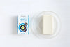 Clearspring Luomu Silken Tofu: pakkaus Clearspring-luomutofua avatun tofupalan vieressä läpinäkyvällä lasilautasella, molemmat valkoisella pinnalla; pakkaus on suorakulmainen mattapinnalla, pääosin valkoinen ja sininen, groteskitekstillä "Clearspring organic tofu"; tofu itsessään on murrettu valkoinen ja suorakulmainen sileine reunoin; ei ihmisiä tai toimintaa näkyvissä.