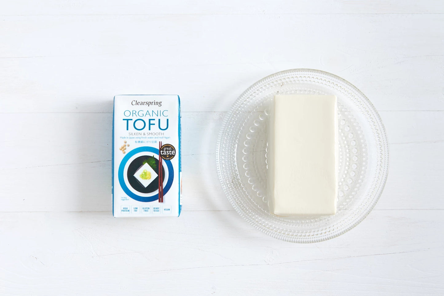 Clearspring Luomu Silken Tofu: pakkaus Clearspring-luomutofua avatun tofupalan vieressä läpinäkyvällä lasilautasella, molemmat valkoisella pinnalla; pakkaus on suorakulmainen mattapinnalla, pääosin valkoinen ja sininen, groteskitekstillä "Clearspring organic tofu"; tofu itsessään on murrettu valkoinen ja suorakulmainen sileine reunoin; ei ihmisiä tai toimintaa näkyvissä.