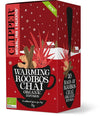 A red box of Clipper Warming Rooibos Chai Organic Infusion tea, featuring a black teacup with cinnamon sticks, star anise, and red leaves. The packaging is rectangular with a matte finish. White sans-serif text identifies the organic product. Background includes illustrations of stylized snow and houses; no people or hands are visible in the image.