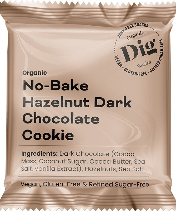 A beige package labeled No-Bake Hazelnut Dark Chocolate Cookie features ingredients and notes that it is vegan, gluten-free, and refined sugar-free. The packaging highlights the presence of rich hazelnuts blended with dark chocolate. The pouch is matte with a rectangular shape and resealable top. Typography varies in size and weight, all clearly legible on the front. The product is photographed against a plain white background without people or objects present.