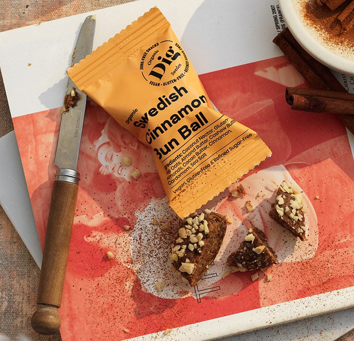 Dig Luomu Kanelipullapallo; osittain avattu Swedish Cinnamon Bun Ball -välipala lepää valkoisella keramiikkalautasella ruostumattomasta teräksestä valmistetun veitsen kera; pakkaus on kirkasta muovia; pyöreä välipala on kullanruskea, siinä näkyy siemeniä tai pilkottuja pähkinöitä; lautasella myös murusia, kokonaisia ja pilkottuja pähkinöitä sekä kolme kanelitankoa, ruskeaa kanelijauhetta ripoteltuna lautasen pinnalle; ei ihmisiä tai käsiä näkyvissä.