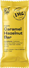 Dig Luomu Karamelli-Hasselpähkinäpatukka; keltainen patukan kääre, merkitty Organic Caramel Hazelnut Bar, korostaa vegaanisuutta, gluteenittomuutta ja puhdistetusta sokerista vapaata koostumusta; superfood-ainesosina cashewpähkinät, hasselpähkinät, taatelit, kookospähkinänektari, merisuola ja vaniljauute; kääre on suorakulmainen mattapinnalla, pääosin lihavaa groteskitypografiaa; ei ihmisiä tai käsiä kuvassa; tausta valkoinen studioasetuksessa, tuote keskellä kuvaa.