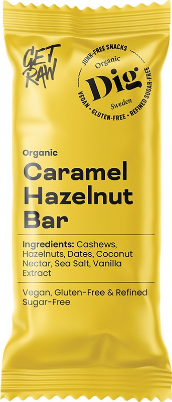 Dig Luomu Karamelli-Hasselpähkinäpatukka; keltainen patukan kääre, merkitty Organic Caramel Hazelnut Bar, korostaa vegaanisuutta, gluteenittomuutta ja puhdistetusta sokerista vapaata koostumusta; superfood-ainesosina cashewpähkinät, hasselpähkinät, taatelit, kookospähkinänektari, merisuola ja vaniljauute; kääre on suorakulmainen mattapinnalla, pääosin lihavaa groteskitypografiaa; ei ihmisiä tai käsiä kuvassa; tausta valkoinen studioasetuksessa, tuote keskellä kuvaa.