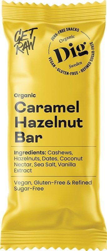 Dig Luomu Karamelli-Hasselpähkinäpatukka; keltainen patukan kääre, merkitty Organic Caramel Hazelnut Bar, korostaa vegaanisuutta, gluteenittomuutta ja puhdistetusta sokerista vapaata koostumusta; superfood-ainesosina cashewpähkinät, hasselpähkinät, taatelit, kookospähkinänektari, merisuola ja vaniljauute; kääre on suorakulmainen mattapinnalla, pääosin lihavaa groteskitypografiaa; ei ihmisiä tai käsiä kuvassa; tausta valkoinen studioasetuksessa, tuote keskellä kuvaa.