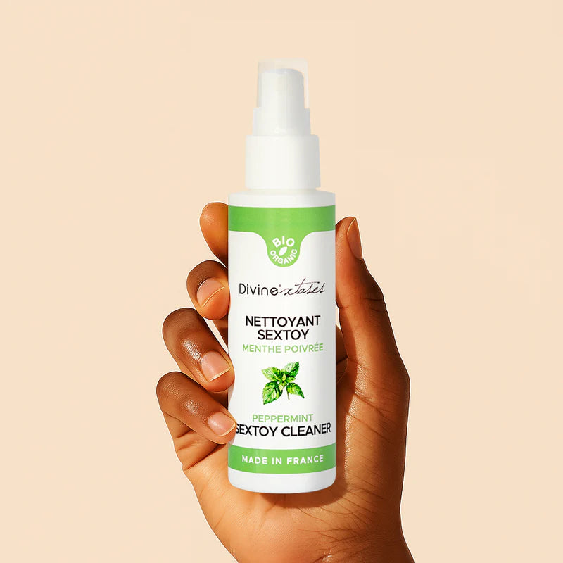 DivineXtases Seksilelujen Puhdistusaine; käsi pitelee valkoista muovista suihkepulloa vihreällä korkilla ja kiiltopinnalla; etiketissä lukee ”Divine Tentation Peppermint Sextoy Cleaner” selkeällä nykyaikaisella mustalla kirjasimella yhdessä minttulehtikuvituksen kanssa; tausta tasainen beige; tuote korostaa raikasta piparmintun tuoksua.