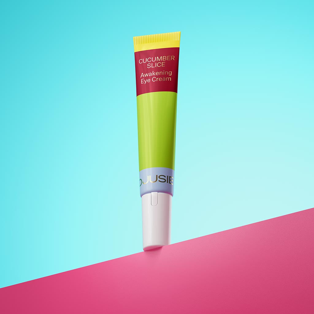 Djusie Cucumber Slice Awakening Eye Cream; tuubi pystyssä pinkillä alustalla sinistä taustaa vasten, vihreä pakkaus keltaisella korkilla ja valkoisella pohjaosalla; etiketissä selkeästi näkyvä tuotenimi groteskifontilla; tuubin pinta vaikuttaa mattapintaiselta; kuvassa ei ihmisiä tai toimintaa.