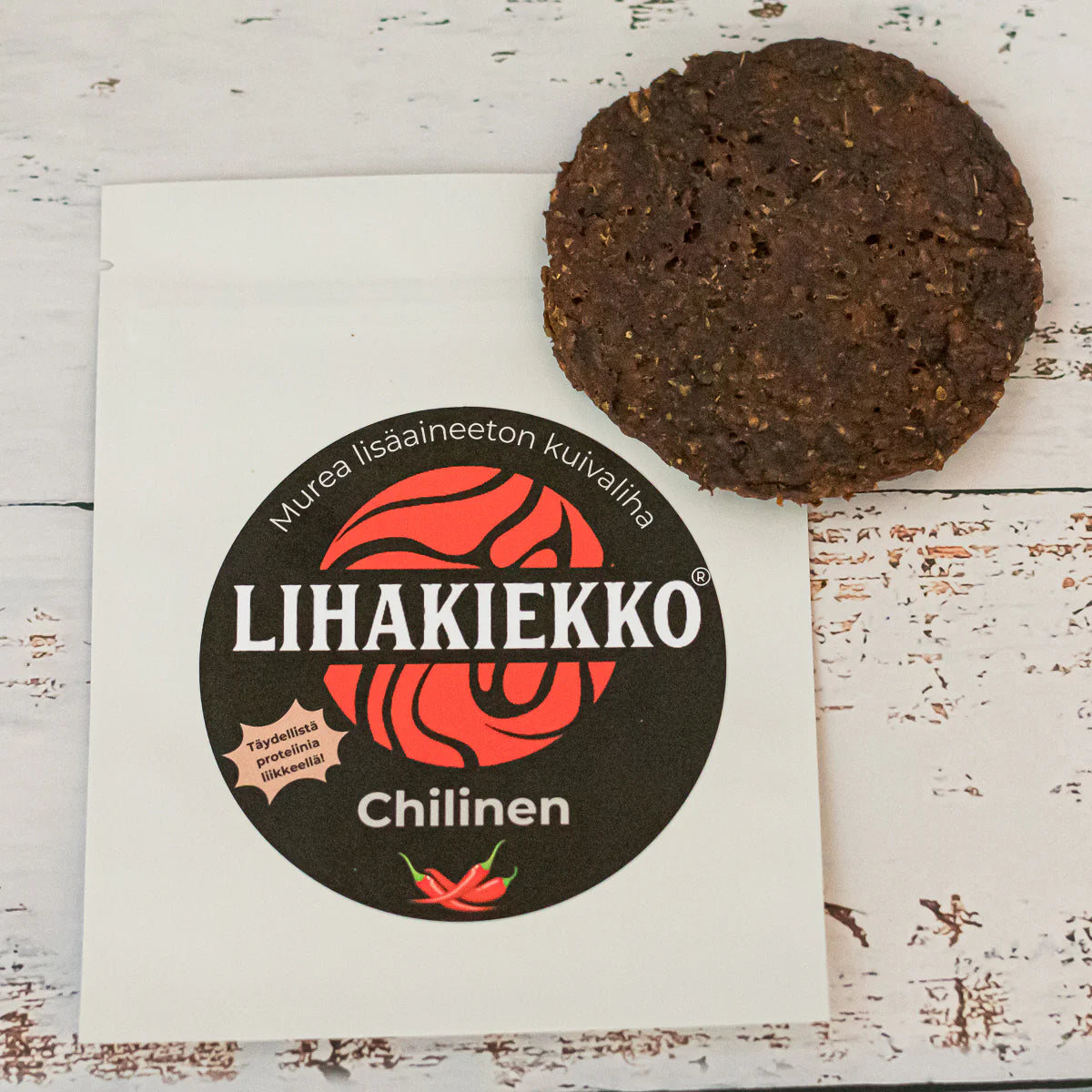 Ellin Lihakiekko Chilinen; pyöreä, tumma kuivalihanaksu lepää osittain mattavalkoisella pussilla, jossa teksti ”Lihakiekko Chilinen” lihavoiduin kirjaimin, punaisia chilipaprikakuvituksia ja lisää suomenkielistä tekstiä; tuote rustiikkisella, valkoiseksi maalatulla puupinnalla; ei ihmisiä tai käsiä näkyvissä kuvassa.