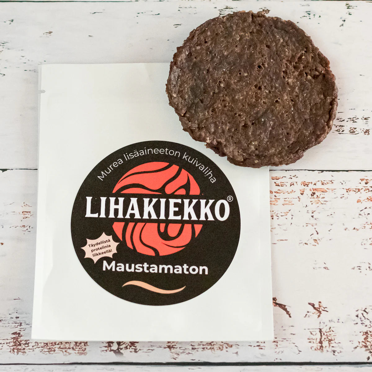 Ellin Lihakiekko Maustamaton; pyöreä pala kuivalihaa valkoisella puupinnalla valkoisen pussin vieressä, tekstillä ”LIHAKIEKKO Maustamaton” ja lihavoitu punainen ja musta logo; kuivaliha tummanruskeaa mattapintaisena; pussi suorakaiteen muotoinen, kiiltäväpintainen ja pääosin valkoinen, etikettitekstissä tikkukirjasinta mustana ja punaisena; ei ihmisiä tai käsiä tässä tuotekeskeisessä kuvassa.