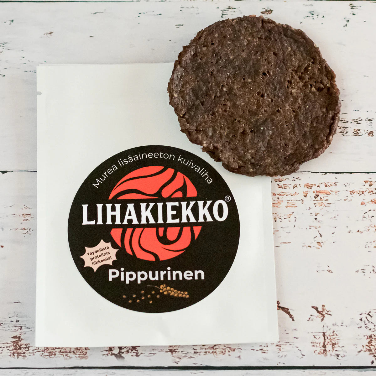 Ellin Lihakiekko Pippurinen; pyöreä kuivattu lihakiekko lepää valkoisella rustiikkisella pinnalla valkoisen pakkauksensa vieressä, jossa lukee ”LIHAKIEKKO Pippurinen”, kuva pippurinsiemenistä ja korostettu maukas pippurin maku; pakkaus mattavalkoinen yksinkertaisella suorakaiteen muodolla ja selkeällä tuotekuvalla; tausta tasainen, ei ihmisiä tai esineitä näkyvissä; kuva otettu ylhäältä.