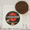 Ellin Lihakiekko Pizzamausteinen; pyöreä, tumma kuivalihapihvi lepää mattavalkoisella pussilla tekstillä ”Lihakiekko” lihavoiduin kirjaimin, vieressä punainen pyörre ja tyylitelty pizzaviipalekuvitus; pakkaus rustiikkisella, kulahtaneella valkoisella puupinnalla; kaikki näkyvä teksti suomeksi; ei ihmisiä tai toimintaa tässä lähikuvassa ylhäältä.