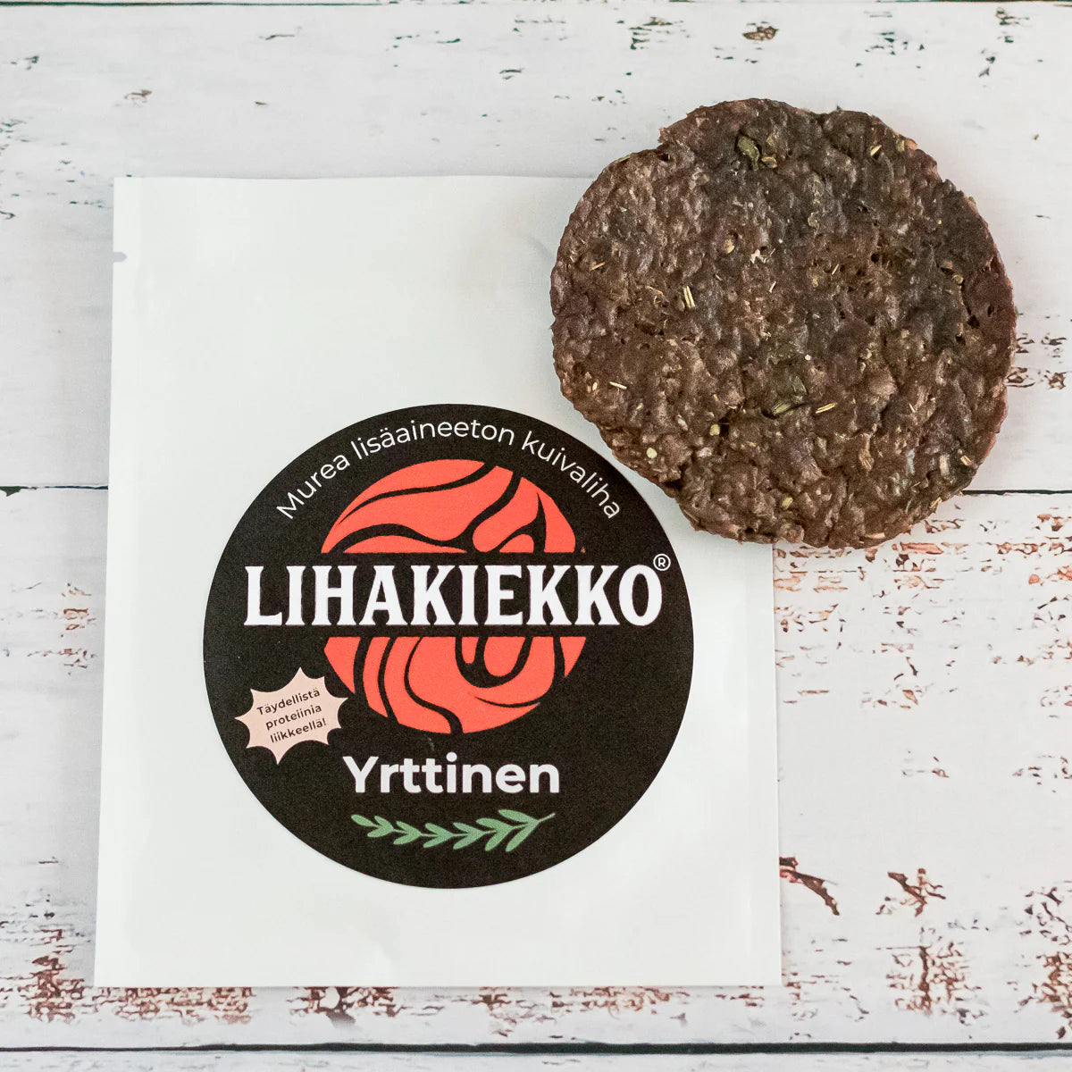 Ellin Lihakiekko Yrttinen; pyöreä, ruskea kuivalihapihvi valkoisen pakkauksensa vieressä, tekstillä ”Lihakiekko Yrttinen” vihrein ja punaisin grafiikoin sekä rustiikkisin yrttiaiheisin tehostein, rustiikkisella valkoisella puupinnalla; pakkaus mattapintainen suorakaiteen muotoinen, lihavoitua tikkukirjasinta tuotenimelle ja värillisiä kasvikuvituksia; ei ihmisiä tai käsiä näkyvissä.