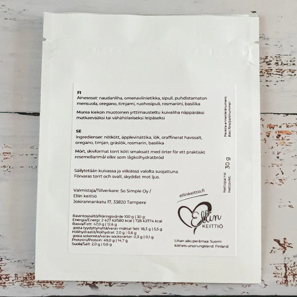 Ellin Lihakiekko Yrttinen; valkoinen elintarvikepussi suomenkielisin tekstein ainesosista ja ravintotiedoista; materiaali mattamuovia, suorakaiteen muotoinen saumatuin reunoin; painettu logo ”Elliin Keittio” selkeällä tikkukirjasimella keskellä etusivua; kääntöpuolella mustia painettuja tekstilohkoja; pussi lievästi kuluneella valkoisella puupöydällä tasaisessa luonnonvalossa ylhäältä kuvattuna.