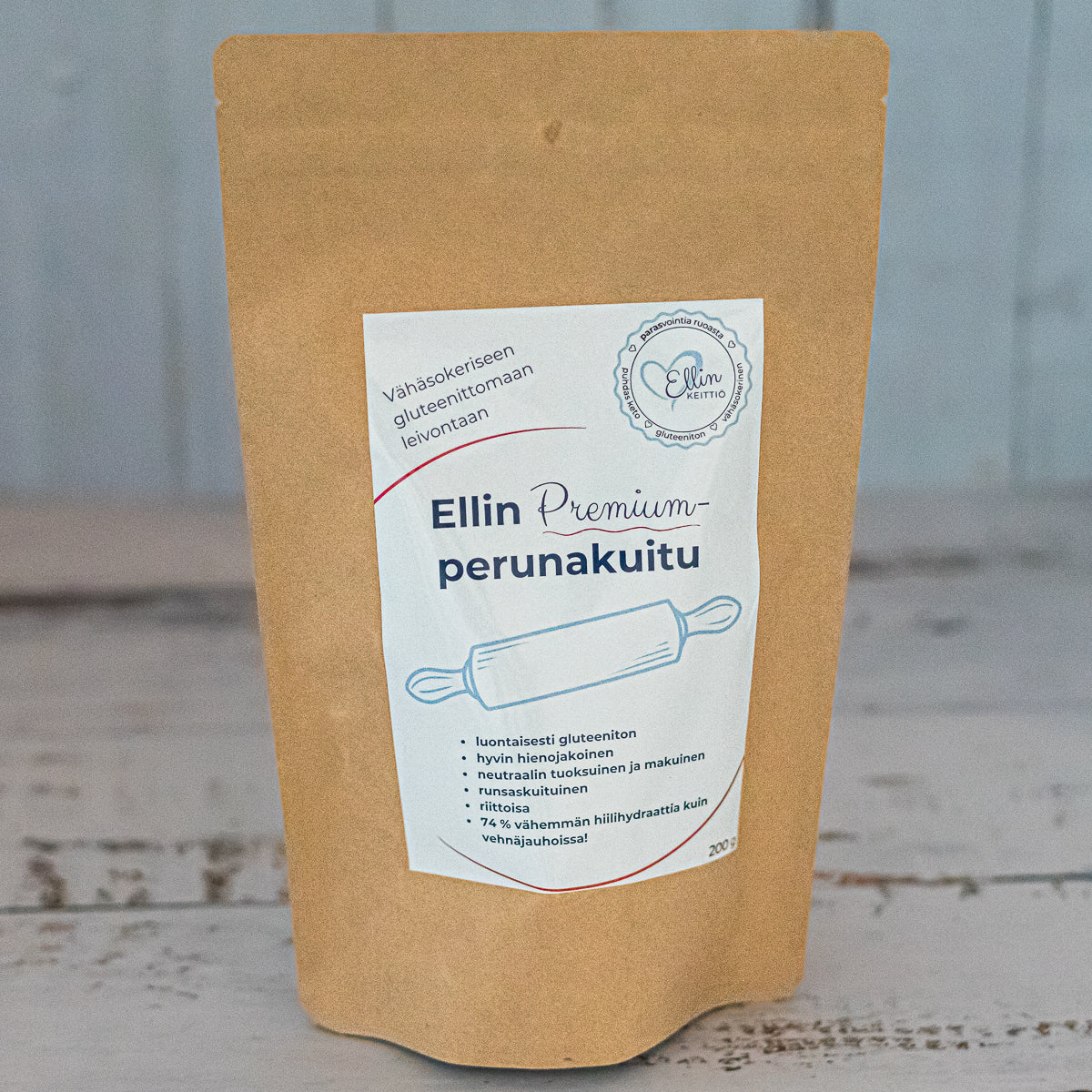 Ellin Premium Perunakuitu; ruskea paperipakkaus, jossa lukee Ellin Premium perunakuitu, leivontaan liittyvää suomenkielistä tekstiä ja ravintokuitutietoa, sijaitsee vaalealla puupinnalla epätarkan taustan edessä; pakkaus on mattaruskea, suorakulmainen ja litteäpohjainen, painettua tummaa groteskitekstiä; ei ihmisiä tai käsiä näkyvissä; kuva otettu pöydän tasolta suoraan edestä.