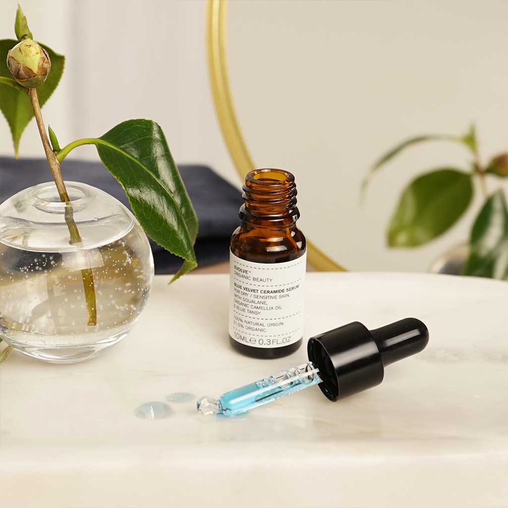 Evolve Blue Velvet Ceramide Serum; pieni ruskea apteekkityylinen pullo etiketillä avattuna marmoripinnalla, vieressä sinistä seerumia sisältävä tiputin; taustalla pyöreä lasimaljakko lehtevine kasveineen; lieriön muotoinen, läpinäkymätöntä meripihkanvärisen lasia mattapintaisena, valkoinen suorakulmainen etiketti groteskitekstillä; tiputin mustalla yläosalla ja kirkkaalla varrella; ei ihmisiä tai käsiä; pehmeästi valaistu marmoripinta.