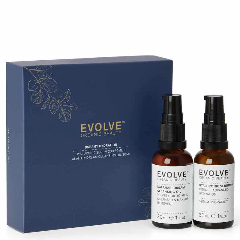 Evolve Dreamy Hydration; kaksi ruskeaa pumppupulloa tekstillä EVOLVE seisoo sinisen laatikon vieressä, jossa lukee EVOLVE ORGANIC BEAUTY Dreamy Hydration; pakkauksessa kulta- ja valkoista tekstiä siistillä groteskifontilla, laatikossa minimalistisia lehtikuvituksia; molemmat pullot mattapintaisia lieriömäisiä muotoja mustin pumpuin; neutraali tausta, ei ihmisiä tai lisärekvisiittaa; suoraan edestä kuvattu tuotekuva.