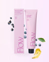 FLOW Cosmetics Balm to Milk Puhdistusvoide: pinkki tuubi ja yhteensopiva rasia tekstillä "Flow Balm to Milk Cleanser" seisovat pystyssä mustaherukoiden, mustikoiden, vihreän lehden ja keltaisen balsamiraidan ympäröiminä vaaleanpinkillä pohjalla; pakkaus on mattapintainen, tuubi pehmeää muovia ja rasia suorakulmainen kartonkia; yksinkertainen valkoinen teksti tuotemerkinnässä; ei ihmisiä tai käsiä näkyvissä, kuva on ylhäältä otettu asetelma.