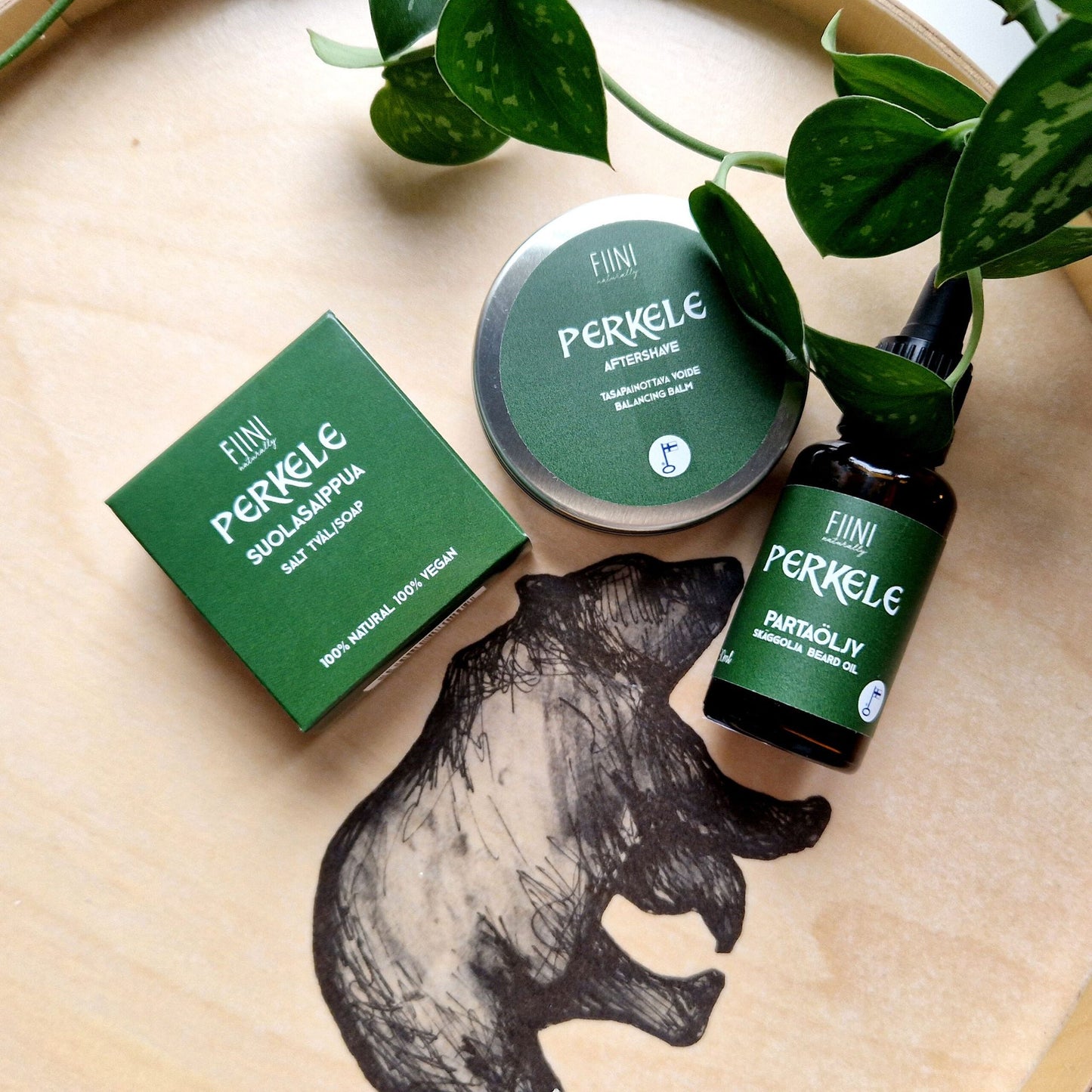 Green-labeled beard care products on a wooden tray with a bear illustration, surrounded by fresh plant leaves for a natural touch. Multiple green containers with matte surfaces and cylindrical or rectangular shapes are arranged on a brown wooden tray. Labels feature simple white sans-serif text and a small bear illustration. No people or hands visible. Background is plain white. Close-up product shot.