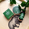 Three green-labeled Finnish skincare products are arranged on a wooden tray featuring an illustration of a bear, beside a lush green leafy plant. The selection includes natural soap, aftershave balm, and beard oil. Labels have simple sans-serif fonts and some Finnish text. Bottles and box shapes include cylindrical glass with matte finishes and a rectangular cardboard box. No people or hands present.