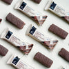Foodin Clean & Real Protein Bar Crispy Chocolate Bliss; suklaapatukoita, sekä kääreettöminä että pakkauksessa merkittynä "Crispy Chocolate Bliss", aseteltuna siistiin, vinoittaiseen kuvioon vaalealle taustalle; suorakulmaiset patukat tekstuurisella suklaapäällysteellä puolikiiltävällä pinnalla; pakkaus suorakulmainen näkyvällä typografisella etikettitekstillä; ei ihmisiä eikä käsiä näkyvissä; kuva ylhäältä.